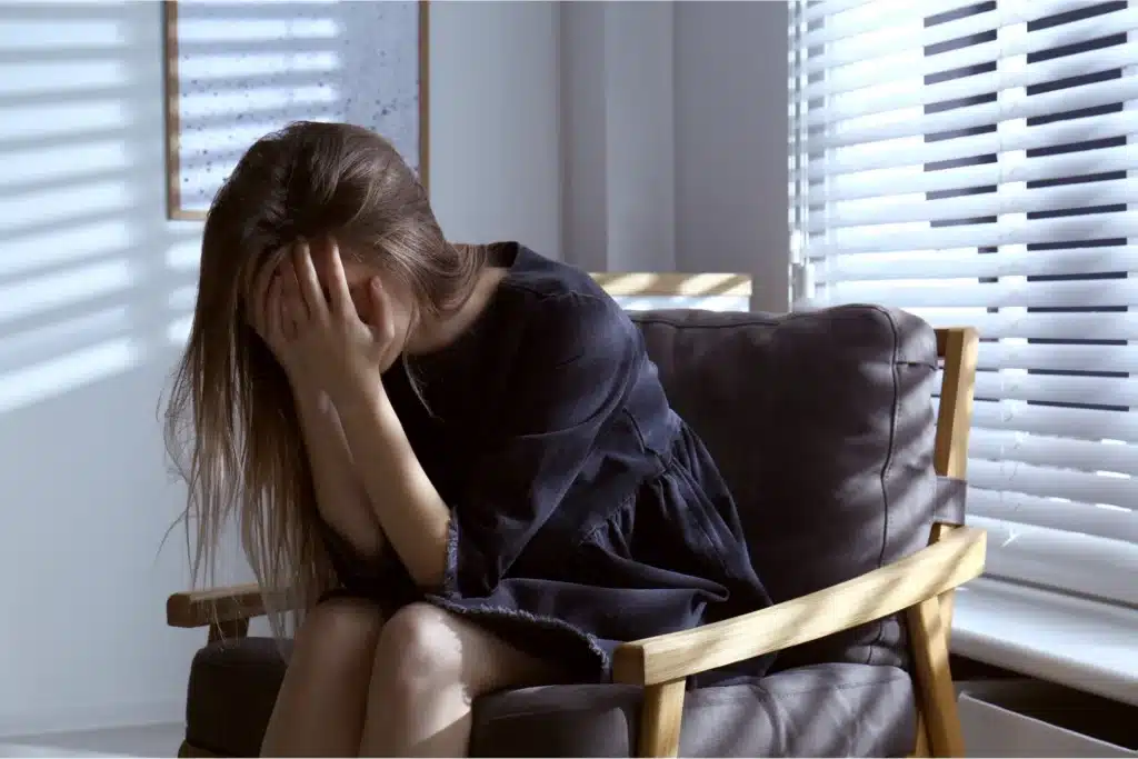 Person sitting in an armchair with head in hands, appearing distressed in a sunlit room.