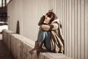 A young woman sitting alone outdoors, looking distressed, reflecting the emotional warning signs of a substance use disorder.
